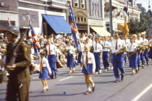 Historische optocht met jeugdige wandelaars in blauwe broeken, witte overhemden en oranje stropdassen en accenten, begeleid door een militair.