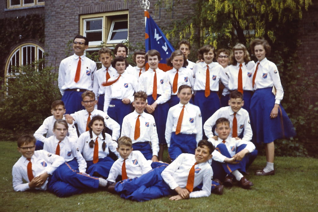 Groepsfoto van jonge wandelaars in blauw-witte uniformen met een verenigings vlag, buiten op het gras.